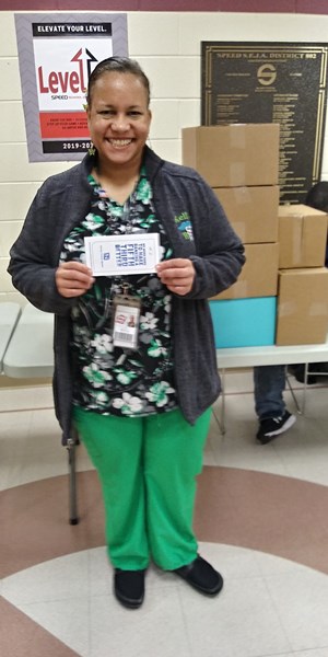 A smiling woman in scrubs holds a card while standing in front of a table with boxes in a community center setting.