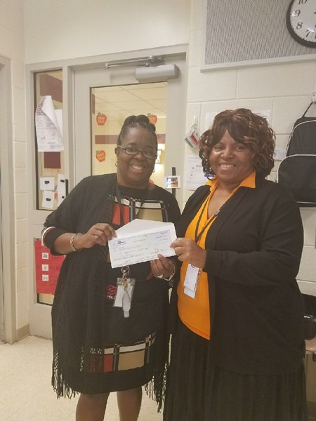 Two women are standing in an office setting, smiling and holding a large check together, with various office items visible in the background.