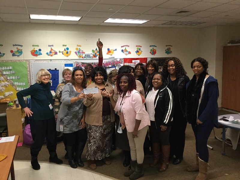 A group of diverse women stands together in a classroom, smiling and posing for the camera, with educational materials visible in the background.