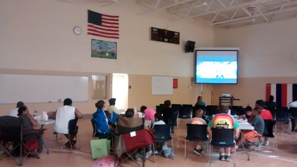 A group of people seated in a classroom setting, facing a projector screen, with an American flag and a clock visible on the wall.