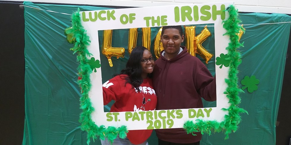 A man and a woman pose together in front of a festive St. Patrick's Day backdrop, framed by a decorative border that reads 'Luck of the Irish' and features green leaves.