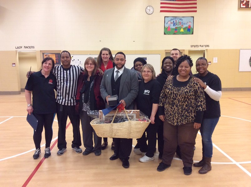 A group of diverse individuals, including a referee and a man in a suit holding a basket, pose together in a gymnasium with an American flag in the background.