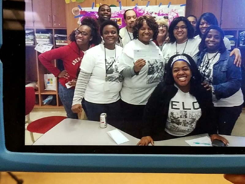 A group of diverse individuals smiling and posing together in a classroom setting, wearing matching white t-shirts with a graphic design, with a festive background.