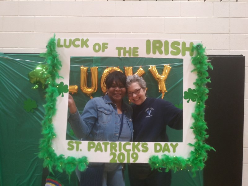 Two people pose together in front of a festive St. Patrick's Day photo frame decorated with green leaves and gold lettering that reads 'Luck of the Irish' and 'Lucky'.