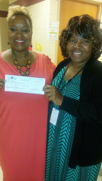 Two women are smiling and holding a large check in a hallway setting.
