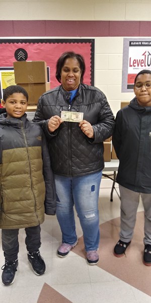 A woman holding a dollar bill stands between two boys in a school setting, with boxes in the background.