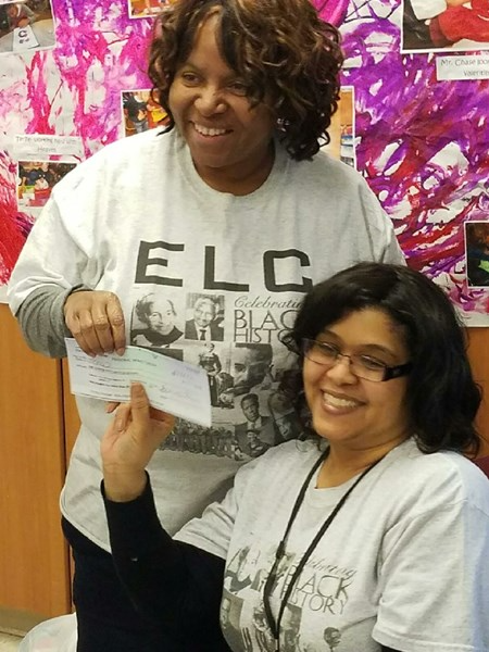 Two women are smiling and posing together, one holding a check while the other sits beside her, both wearing matching gray t-shirts with printed designs.