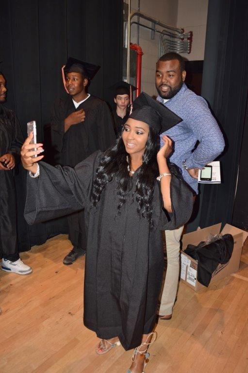 A graduate in a black gown and cap takes a selfie while posing with others in the background, who are also dressed in graduation attire.