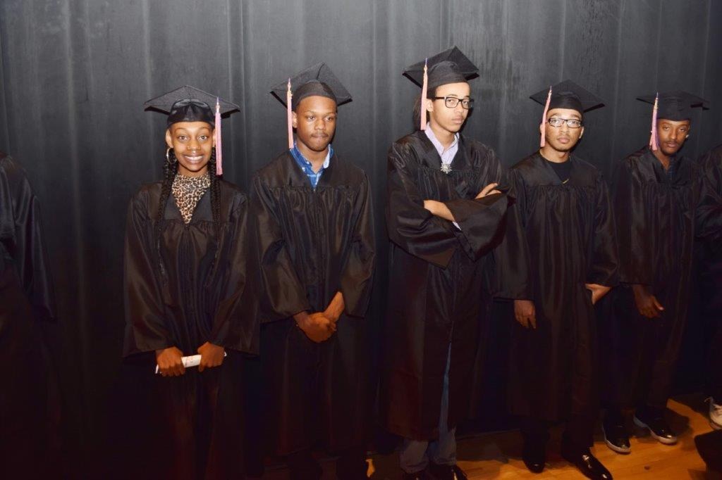 A group of five graduates wearing black caps and gowns stands in a line against a dark backdrop, with expressions ranging from smiles to serious looks.