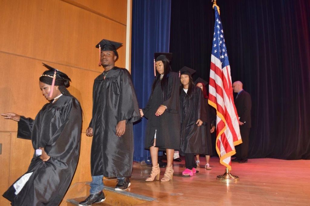 A group of graduates in black gowns and caps walks across a stage, with an American flag in the background.