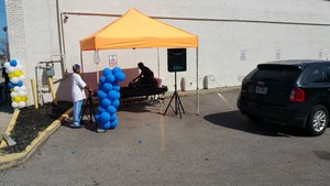 An outdoor event setup featuring an orange tent, a person seated at a table, and a decorative column of blue and yellow balloons, with a parked car nearby.