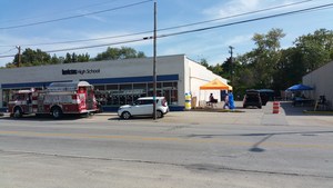 A street view of a building with a sign that reads 'Batteries Plus Bulbs,' featuring a fire truck parked in front and tents set up nearby, indicating a community event or gathering.
