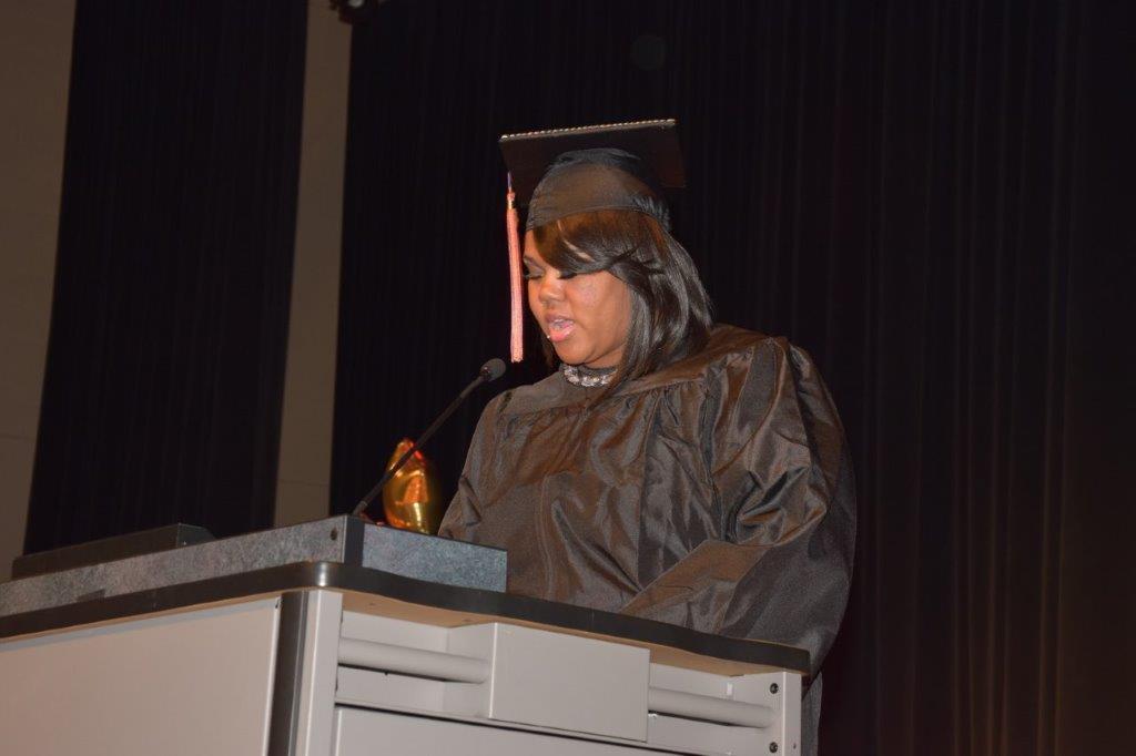 A person wearing a graduation gown and cap stands at a podium, speaking into a microphone during a ceremony.