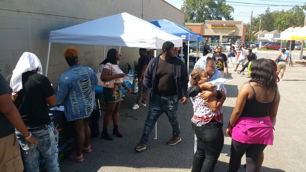 A group of people gathered outdoors near tents, some interacting while others are in line, with a sunny atmosphere and a variety of activities happening in the background.