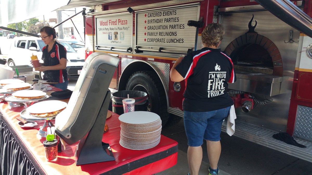 Two people are working at a food truck that resembles a fire truck, with a menu displayed on the side and plates set up on a table in front.