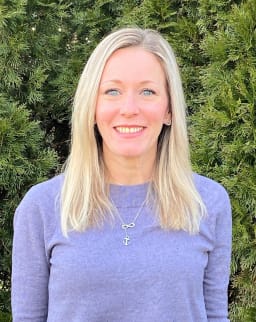 A woman with long blonde hair stands in front of a green bush, wearing a lavender sweater and silver jewelry.