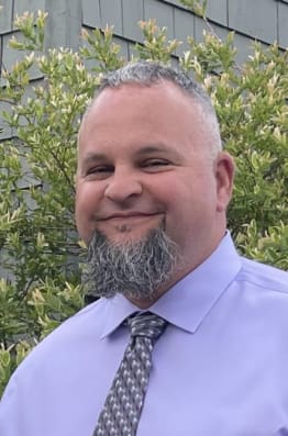 A man in a light purple shirt and patterned tie stands outdoors, with green bushes and a textured wall in the background.