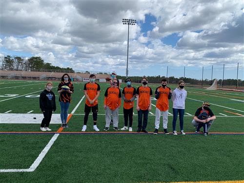 A group of nine students wearing matching orange shirts and masks stands on a football field under a cloudy sky.