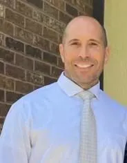 A man in a light blue dress shirt and striped tie stands against a brick wall, exuding a professional demeanor.