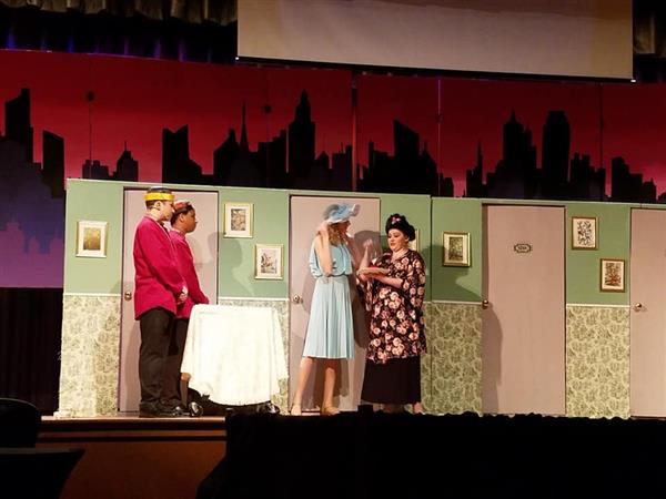 Four actors perform on stage in a colorful set, engaging in a comedic scene with a table and decorated background.