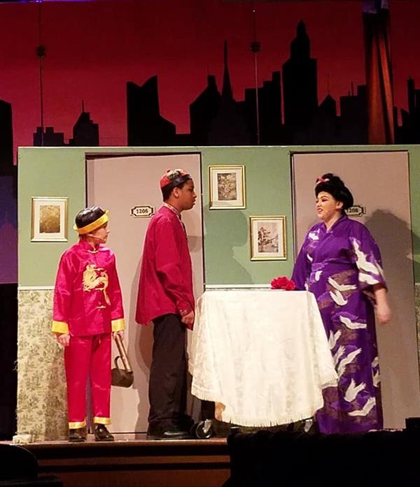 Three actors dressed in colorful traditional costumes stand near two doors, with a table draped in white between them. The backdrop features city silhouettes.