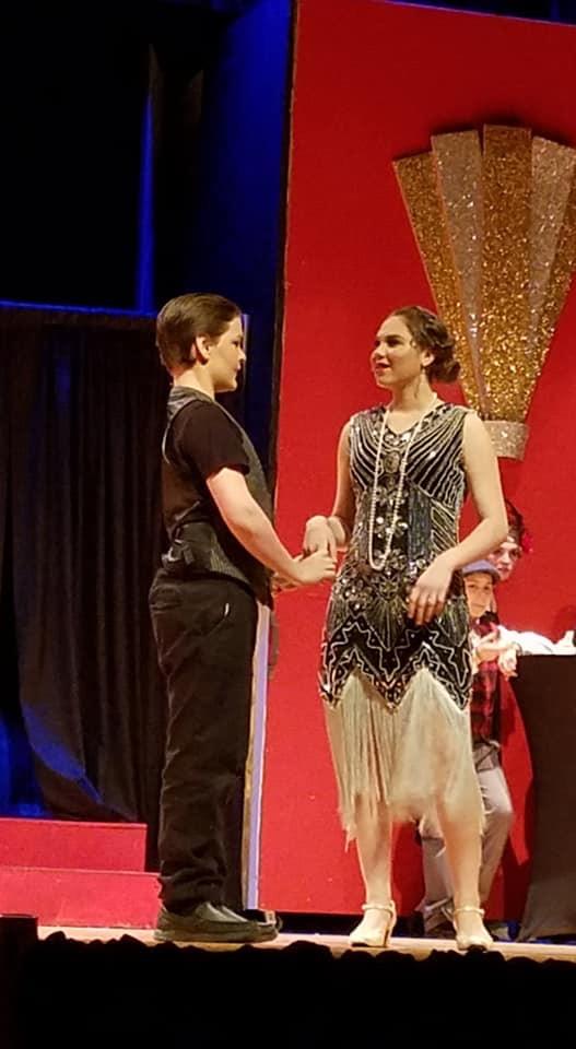 Two performers on stage, dressed in 1920s-inspired outfits, holding hands against a vibrant red and dazzling backdrop.