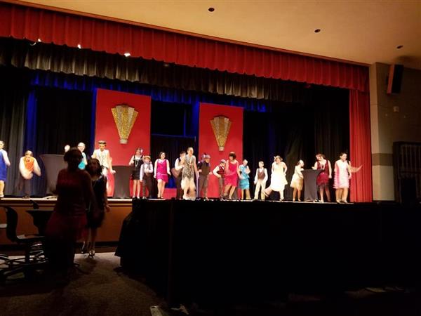 A group of performers in colorful costumes dances on stage, with vibrant red and blue backdrops and audience seating visible in front.