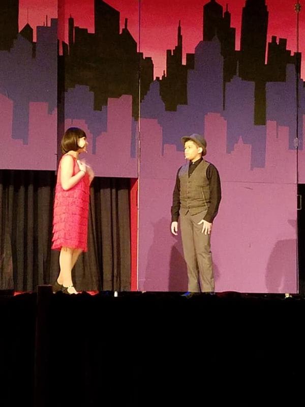 A woman in a pink flapper dress and a man in vintage attire perform on stage, set against a colorful city skyline backdrop.