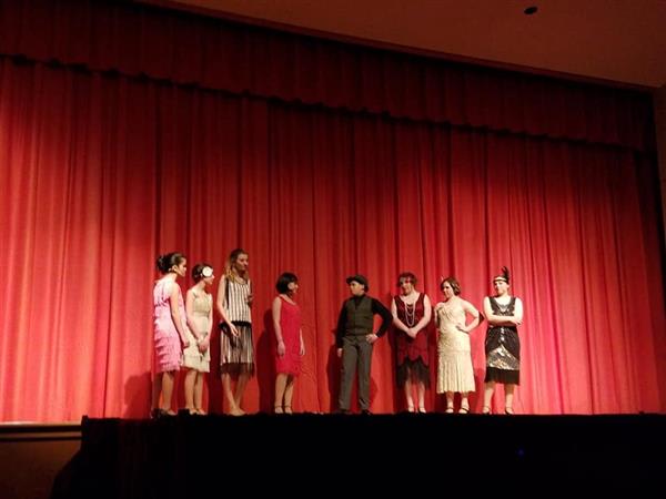 A group of eight performers in vintage 1920s attire stand on stage, with a red curtain backdrop, engaged in a theatrical performance.