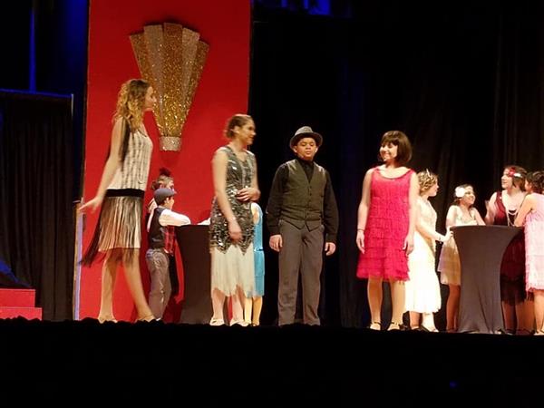 A lively stage performance featuring actors in vintage 1920s costumes, with vibrant colors and elegant set designs.