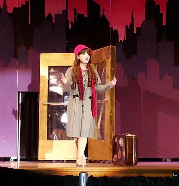 A performer dressed in a gray coat and pink hat stands on stage beside a vintage suitcase and an ornate golden door.