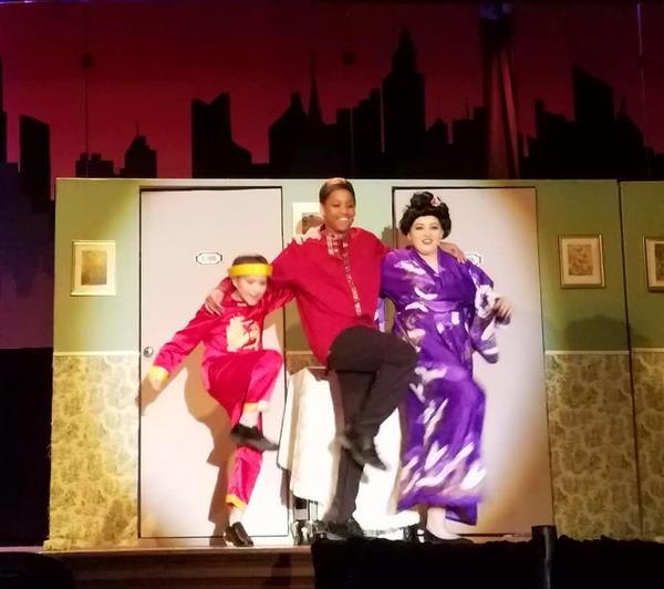 Three performers in vibrant costumes dance on stage, set against a city skyline backdrop, highlighting a lively theatrical scene.
