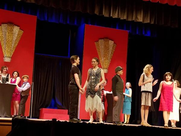 A lively theatrical performance featuring actors in vintage costumes on stage, with vibrant red backdrops and elegant decorations.