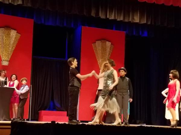 A theatrical performance captures actors in costume on stage, featuring vibrant red backdrops and dramatic lighting.