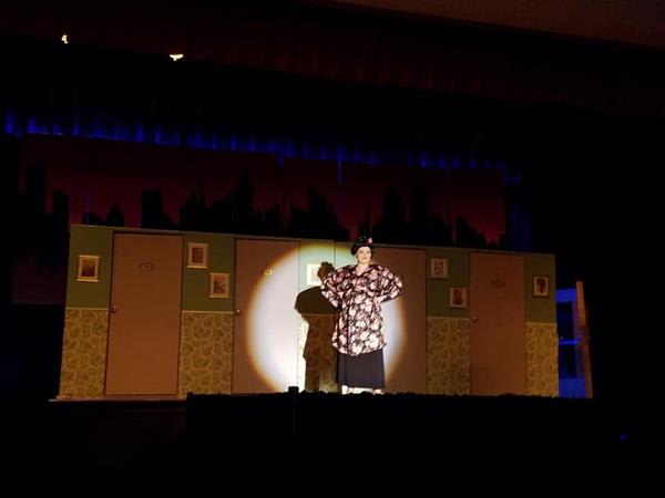 A performer in a floral costume stands under a spotlight on stage, with a backdrop of doors and a city skyline silhouette.