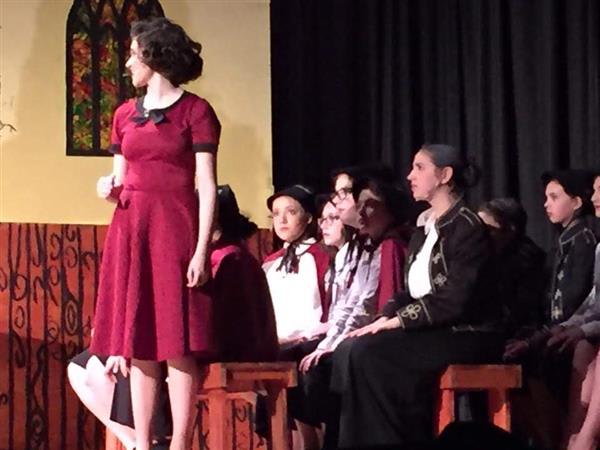 A performer in a red dress stands confidently on stage, surrounded by seated cast members in a theatrical setting.