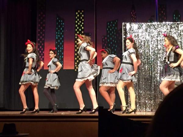 A group of dancers in matching grey dresses and high heels performs on stage, with a sparkling backdrop and city skyline decorations.