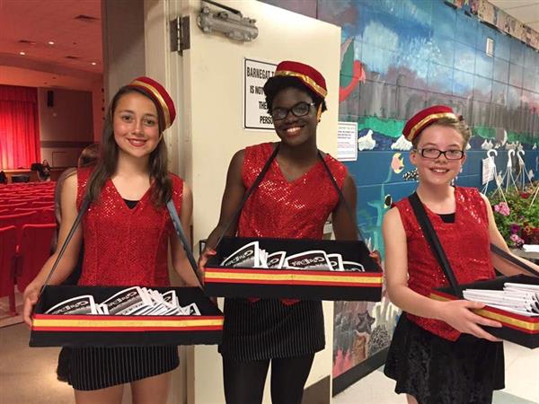 Three performers in sparkly red costumes hold trays of programs in an auditorium with colorful painted walls and red seats.