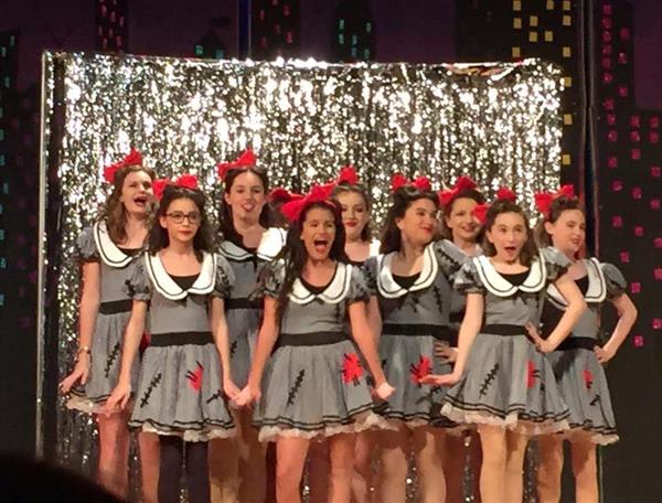 A group of young performers in matching gray dresses with red accents and bows, standing in front of a shiny silver backdrop.
