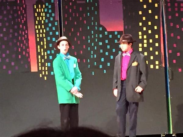 Two performers in colorful costumes stand on stage, with a vibrant city skyline backdrop, engaged in a theatrical performance.