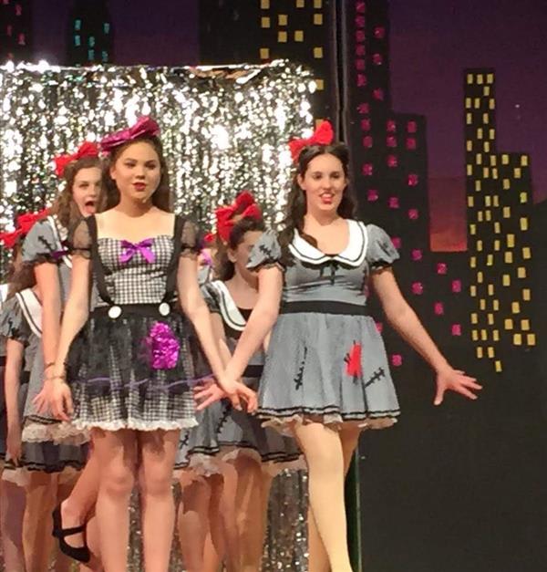 A group of young performers in colorful costumes with vibrant bows, posing on stage in front of a shimmering backdrop.