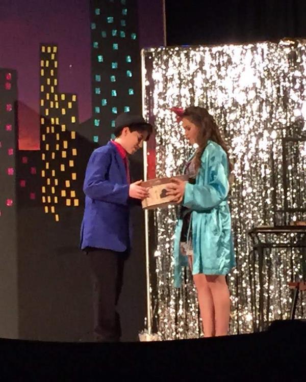 Two performers on stage interact while holding a box, with a colorful city backdrop and sparkling curtain enhancing the scene's theatricality.