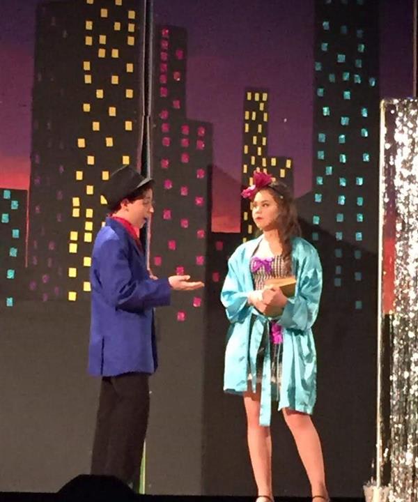 Two performers on stage in colorful costumes, with a vibrant city skyline backdrop, engaging in a dramatic dialogue.