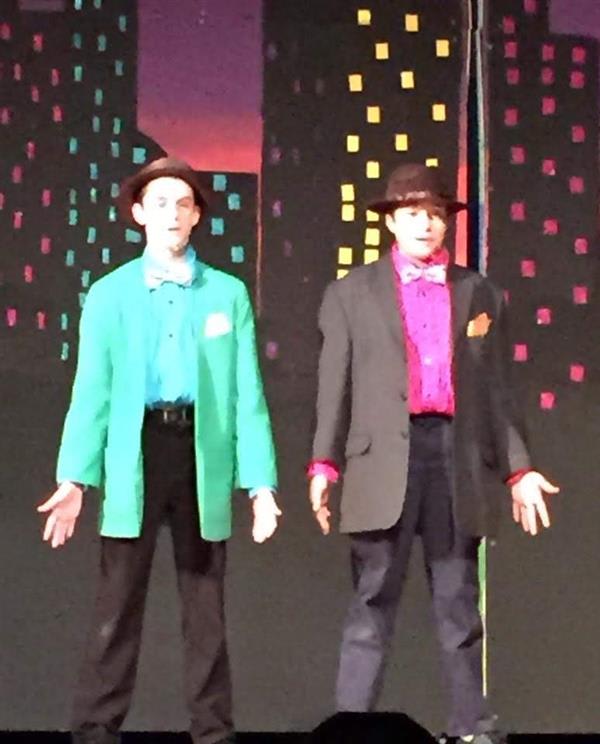 Two performers on stage wearing bright costumes, one in a green jacket and the other in a gray suit, set against a colorful city backdrop.