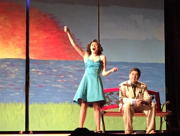 A girl in a blue dress passionately performs on stage while a man in a light suit sits calmly on a bench against a sunset backdrop.