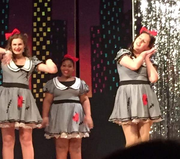Three performers in matching grey dresses with red bows and flower accents are on stage, showcasing a lively dance routine against a colorful backdrop.