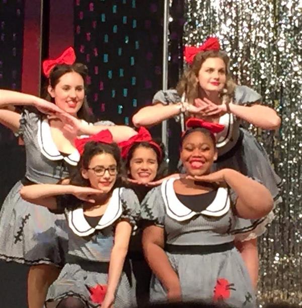 A group of five performers in matching gray dresses with white collars and red bows, posing dramatically on stage with a sparkly backdrop.