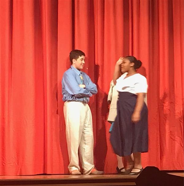 Two students perform on stage against a red curtain; one stands with arms crossed, while the other gestures expressively.