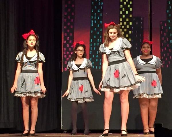 Four girls in matching blue dresses with red bows perform on stage, set against a colorful cityscape backdrop.