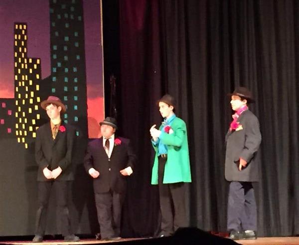 Four performers in suits and hats stand on stage, adorned with colorful roses, against a backdrop of a cityscape at dusk.